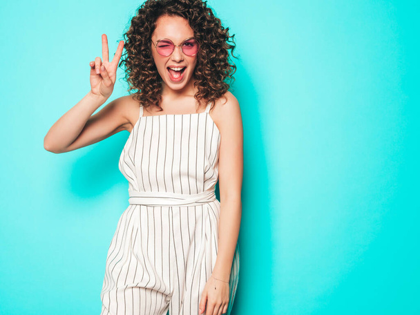Portrait of beautiful smiling model with afro curls hairstyle dressed in summer hipster clothes.Sexy carefree girl posing in studio near blue wall.Trendy funny and positive woman shows peace sign - Foto, Imagem