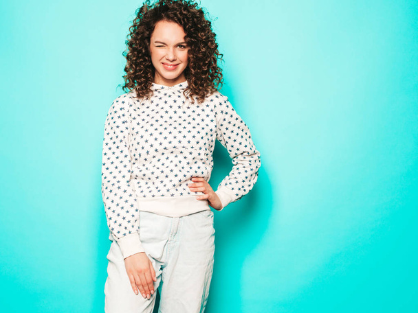 Portrait of beautiful smiling model with afro curls hairstyle dressed in summer hipster clothes.Sexy carefree girl posing in studio near blue wall.Trendy funny and positive woman winking - 写真・画像