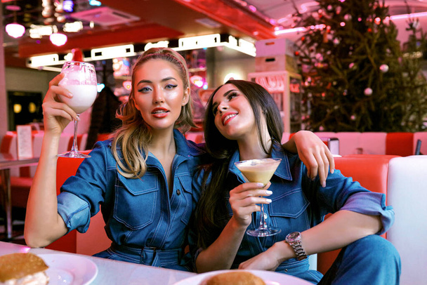 Deux copines élégantes étonnantes se détendre ensemble dans le café. Lumineux néons sur le fond. Elle tenait un verre de cocktail dans sa main. Grandes lèvres avec rouge à lèvres, maquillage et coiffure
. - Photo, image