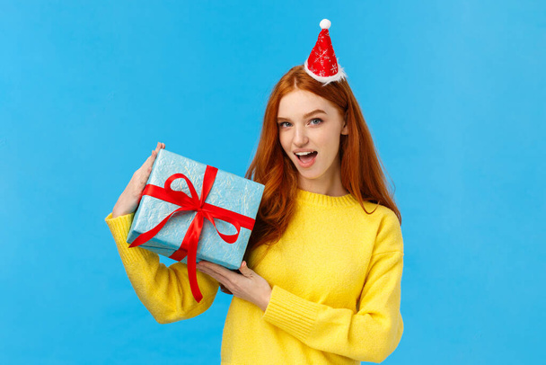Check-out my gift. Sassy and daring good-looking redhead woman show-off, bragging with wrapped christmas present, looking camera happy and cheeky, wearing cute new year hat, blue background - Photo, Image