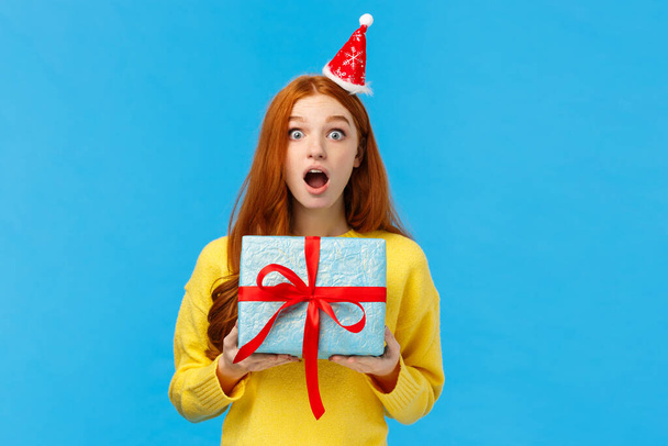 Удивлённая рыжая девушка получает неожиданный подарок на Рождество однокласснице, открытый рот удивлён и молча смотрит в камеру, держа милую синюю упаковку подарка, студийный фон
 - Фото, изображение