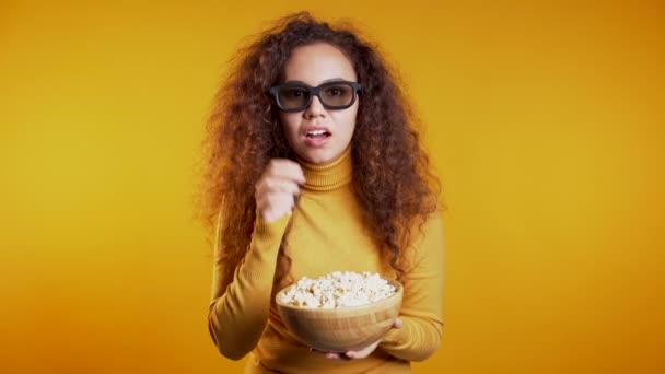 Young woman in 3d glasses watching movie and eating popcorn on yellow studio background.  - 映像、動画