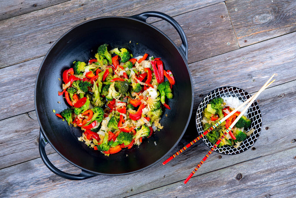 wok with colorful vegetables, bowl of rice and chopsticks on wooden ground - Zdjęcie, obraz
