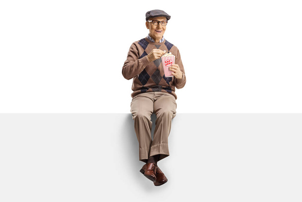Senior man sitting on a panel eating popcorn and miling at camer - Foto, Imagem