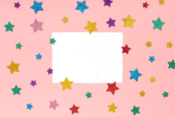 Stars glitters sprinkles in different colors and sizes on pink trendy background with paper card note for text. Festive holiday background. Celebration concept. Mock up, top view, flat lay, copy space - Photo, image