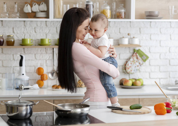 Η νεαρή μαμά αγκαλιάζεται με τον μικρό της γιο στην κουζίνα. - Φωτογραφία, εικόνα