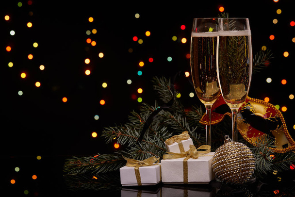 Two glasses with champange, fir tree branch with decoration, gift boxes on a black background with multi-colored lightes of garland.  New year and Christmas. - Photo, Image