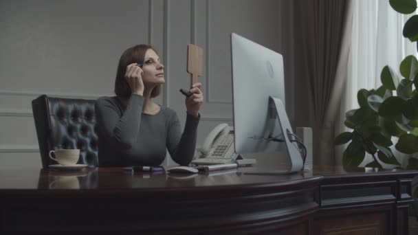 Young businesswoman applying make up looking in mirror at working place. Brunette in business office talking on smartphone. Desk with monitor.  - Filmati, video