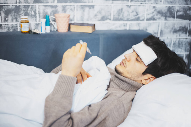 Πορτρέτο του νεαρού άρρωστου άντρα ξαπλωμένος στο κρεβάτι κρατώντας θερμόμετρο στο χέρι σε φόντο άλλων φαρμάκων. - Φωτογραφία, εικόνα