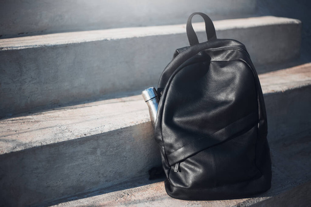 Black backpack with reusable steel thermo water bottle inside on urban stairs. - Фото, изображение
