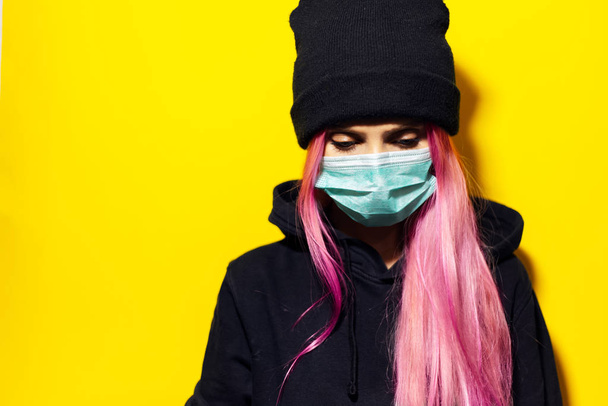 Estúdio retrato de menina com cabelo rosa e olhos azuis, vestindo máscara de gripe médica, vestida com camisola de capuz preto e chapéu de gorro no fundo amarelo
. - Foto, Imagem