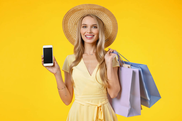 Photo of happy young lady showing display of phone standing isolated over yellow background with shopping bags. - Photo, Image