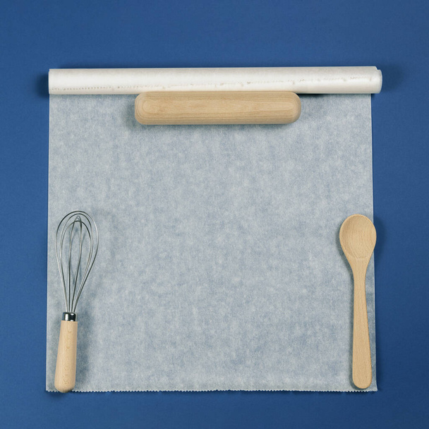 Cuillère en bois, rouleau à pâtisserie et ballon fouettent sur papier pâtissier blanc sur fond bleu. Vue de dessus, plan plat
 - Photo, image