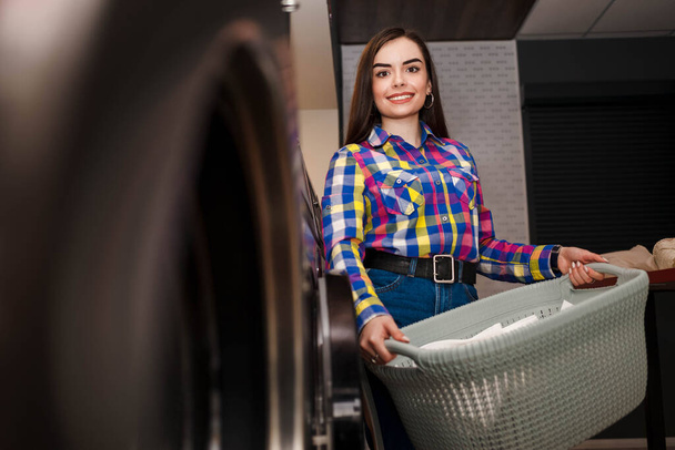 Menina sorridente fica na lavandaria e segura uma cesta de roupa
 - Foto, Imagem