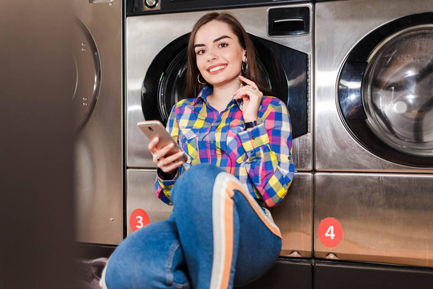 Красива дівчина зі смартфоном в руці слухає музику, що спирається на пральну машину в громадському пральні
 - Фото, зображення