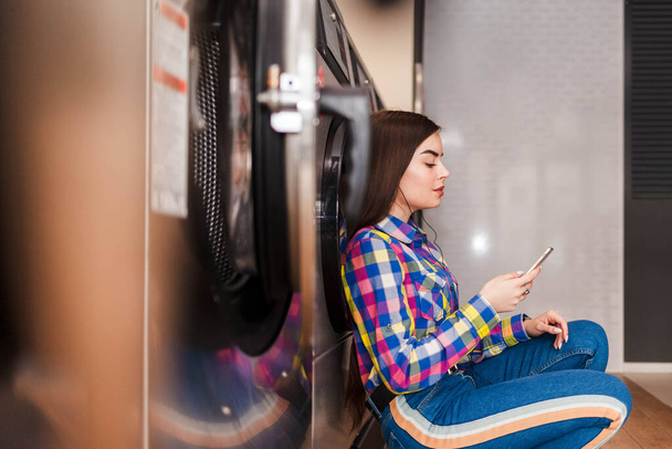 Chica sentada en el suelo en una lavandería y escuchando música en los auriculares. Concepto de proceso de lavandería
 - Foto, imagen