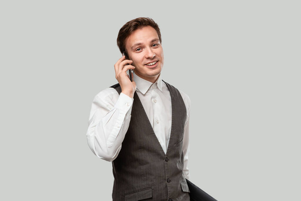 Úspěšný podnikatel hovořící po telefonu drží notebook izolovaný přes šedé pozadí. - Fotografie, Obrázek