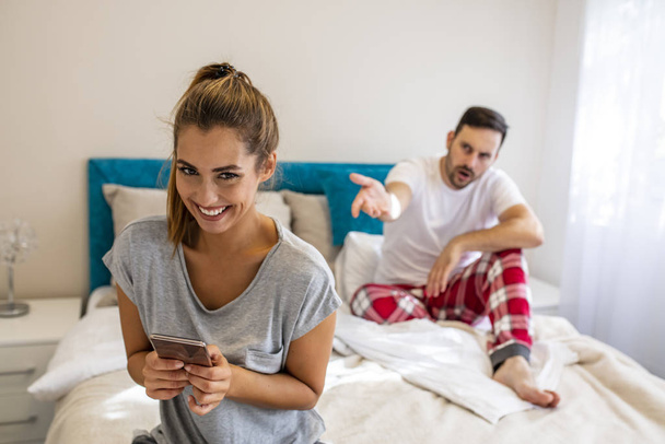 Couple in bed, happy smiling woman turned her back to man, reading message on phone from her lover, worried boyfriend lying next to her, trying to peek at screen. Cheating and infidelity concept - Photo, image