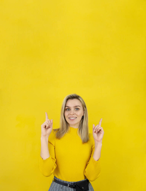 Portrét krásné ženy ve žlutém ukazuje prstem nahoru, usmívá se a dívá se na kameru na žlutém pozadí. Vertikální nápis s mezerou pro kopírování - Fotografie, Obrázek