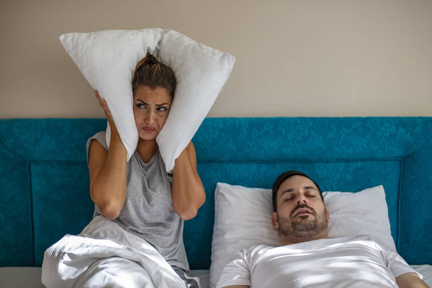 Young couple have problem with man's snoring. Heterosexual couple in bed, man sleeps and snoring with mouth open, while a tired woman irritated by snoring sitting on bed with a pillow on her head. - Photo, Image