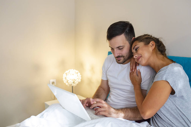 Side view of young couple with digital laptop with blank screen resting on bed at home. Young married couple lying on bed at home and using laptop. Attractive woman embracing husband while using a laptop in bed - Foto, imagen