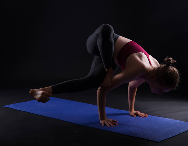 Mujer joven haciendo pose de yoga y asana ashtavakrasana. Chica de fitness disfrutando del yoga en interiores en ropa deportiva sobre fondo negro
. - Foto, imagen