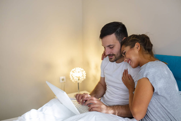 自宅でノートパソコンを使用して幸せなカップル。若いカップルが寝室で楽しんでいます。美しい笑顔の若いカップルの愛の家庭では、ベッドに横たわって、ラップトップコンピュータを見ている。ベッドにノートパソコンがあるカップル - 写真・画像