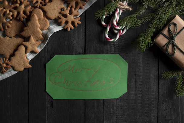 "Directly above view with black wooden table with christmas sweet food, a gift box and a green paper with the message ""Merry Christmas""" - 写真・画像