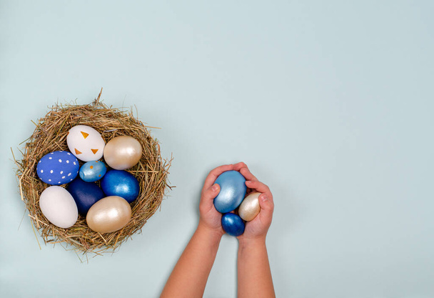 Childrens hands are holding Easter eggs in blue on a blue background with place for copy space - 写真・画像