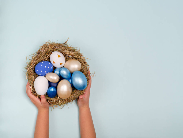 Childrens hands are holding a nest of straw with blue eggs on a blue background with space for copy - Φωτογραφία, εικόνα