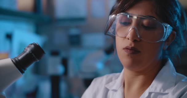Női kutató tudós dolgozik egy laboratóriumban, egy mikroszkóp mellett, fehér kabátot és védő üveget.Kék világítás egy sötét szobában.Közelkép, kézi, lövés Bmpcc 4k. Biokémia - Felvétel, videó