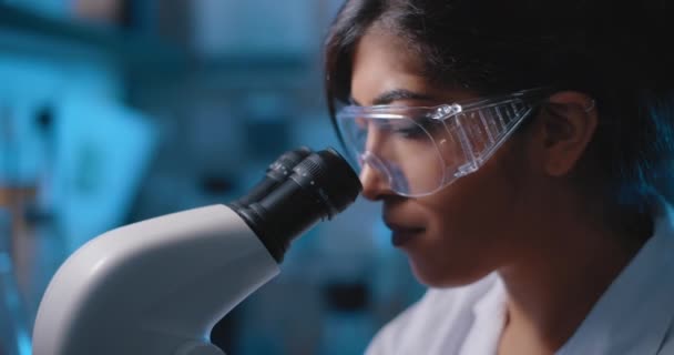 Kobieta naukowiec patrząc w mikroskop, noszenie szkła ochronnego.Niebieskie oświetlenie w ciemnym pokoju laboratoryjnym.Zamknij się, suwak, strzał z Bmpcc 4k.Biochemia, medycyna farmaceutyczna, koncepcja nauki - Materiał filmowy, wideo