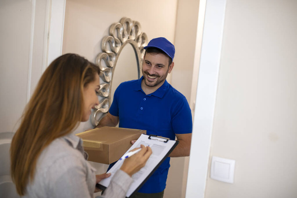 Woman Signing For Package From Courier At Home. Smiling young delivery man holding a cardboard box while beautiful young woman putting signature in clipboard - Foto, imagen
