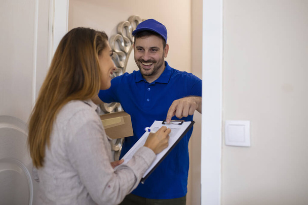 Fast and reliable service. Cheerful young delivery man giving a cardboard box to young woman while standing at the entrance of her apartment. Customer Signing For Delivery From Courier - Фото, зображення