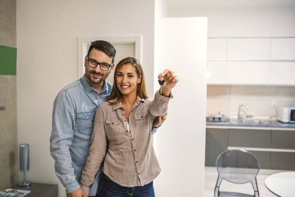 Couple with keys to new home. Cheerful young couple holding keys and smiling while standing in their new house. Happy couple in their new home holding key - Fotoğraf, Görsel