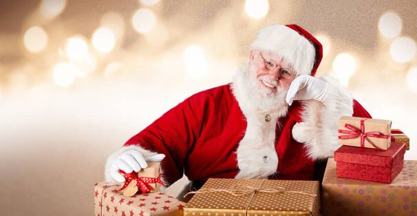 Merry Father Christmas with colorful gifts sitting smiling at the camera against a sparkling bokeh of party lights with copy space in banner format - Foto, Imagem