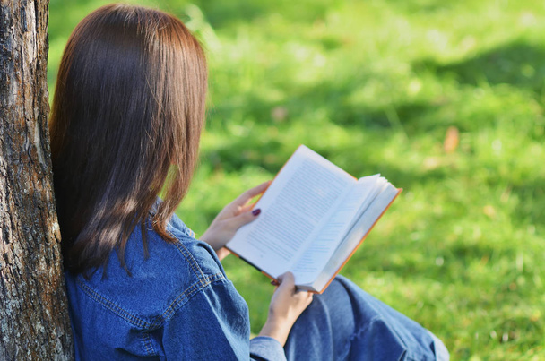 Linda, jovem estudante feliz lendo um livro sentado na grama verde sob uma árvore perto do campus, Universidade, escola, educação. Verão, primavera, parque verde, sol brilhante - Foto, Imagem