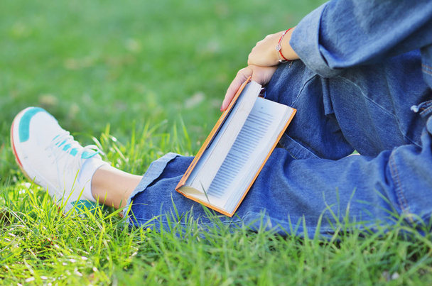 The young girl relaxed while reading an interesting book sitting next to a tree on the green grass - Фото, изображение