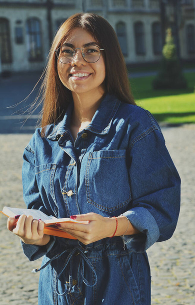 A student in a denim suit stands on the territory of the University and holds an open book in her hands, looks smiling at the camera - Valokuva, kuva