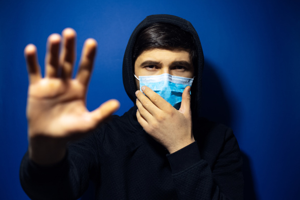 Portrait studio de jeune homme, portant un masque antigrippal médical et un pull à capuche, montrant le geste Stop sur fond de couleur bleue
.  - Photo, image