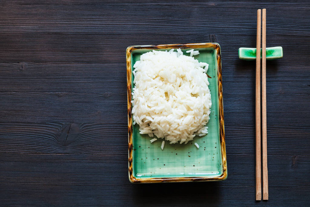 top view of portion of boiled rice on green plate and chopsticks on rest on dark wooden desk with copyspace - Foto, Imagem