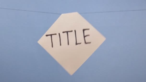Um homem pendura uma folha branca de papel com uma inscrição preta "título" em um fundo azul
 - Filmagem, Vídeo