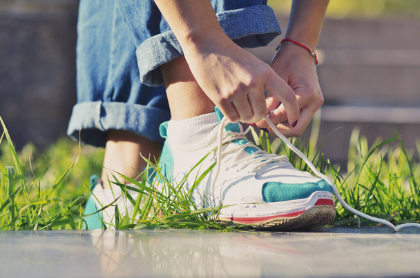 Young girl wearing jeans tying shoelaces on sneakers standing on green grass side view close-up horizontal photo, Sunny day - Foto, afbeelding