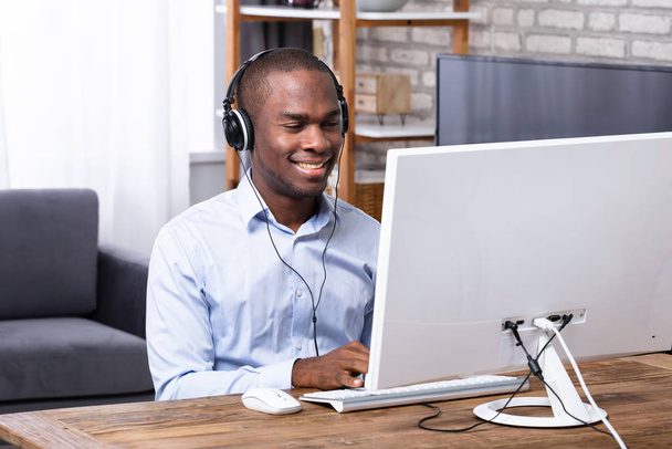 Happy African Man Using Headphone While Working On Computer Over Wooden Desk - Fotografie, Obrázek