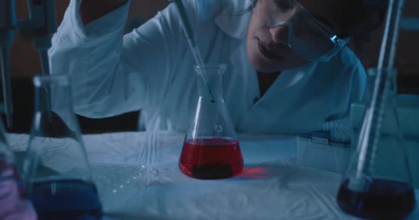 Vrouwelijke onderzoeker voegt een druppel toe in een erlenmeyer met rode vloeistof.Blauwe verlichting in een donkere laboratoriumruimte.Medium, dolly out, slow motion, shot met Bmpcc 4k.Concept: chemie, wetenschap - Video