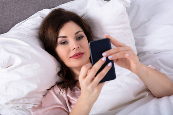 Beautiful Young Woman Lying On Bed Holding Mobile Phone - Foto, Imagen