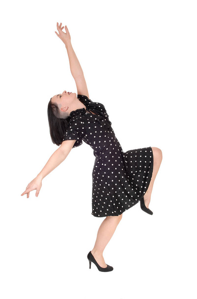 A beautiful young woman dancing in a black pock dot dress with her.arms raised, standing on one leg, isolated for white background. - Foto, Imagen