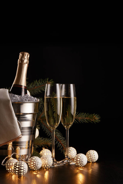 Still life with champagne bottle standing in a bucket with ice, two full champagne flutes, LED lights garland  and Christmas ornaments on a black background. Christmas and New Year celebration - Foto, Imagen