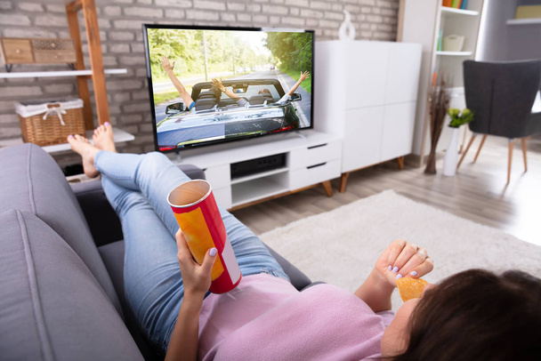 Woman Relaxing On Sofa Eating Potato Chips While Watching Television At Home - Foto, imagen