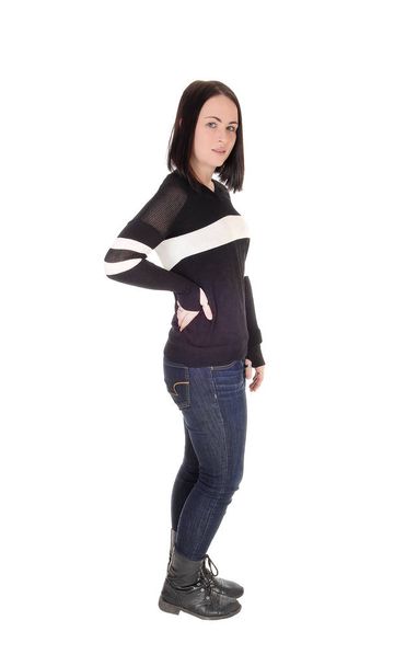 A beautiful young woman standing on white background in jeans and.a sweater with short black hair and one hand on her hip. - 写真・画像